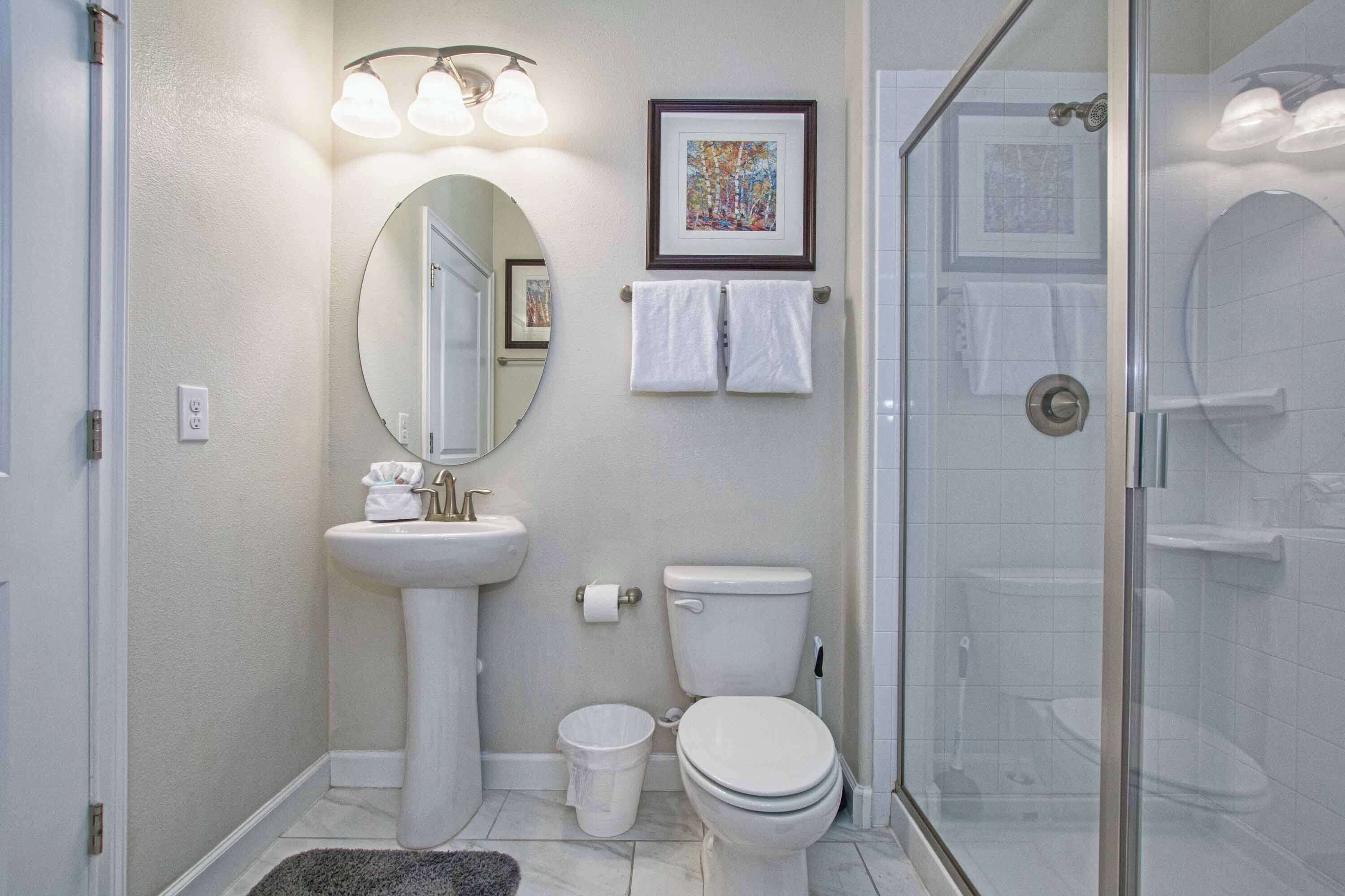 Ein Badezimmer mit einem Standwaschbecken, Toilette und einer Duschkabine. An der Wand hängt ein gerahmtes Bild und ein Handtuchhalter mit zwei Handtüchern.