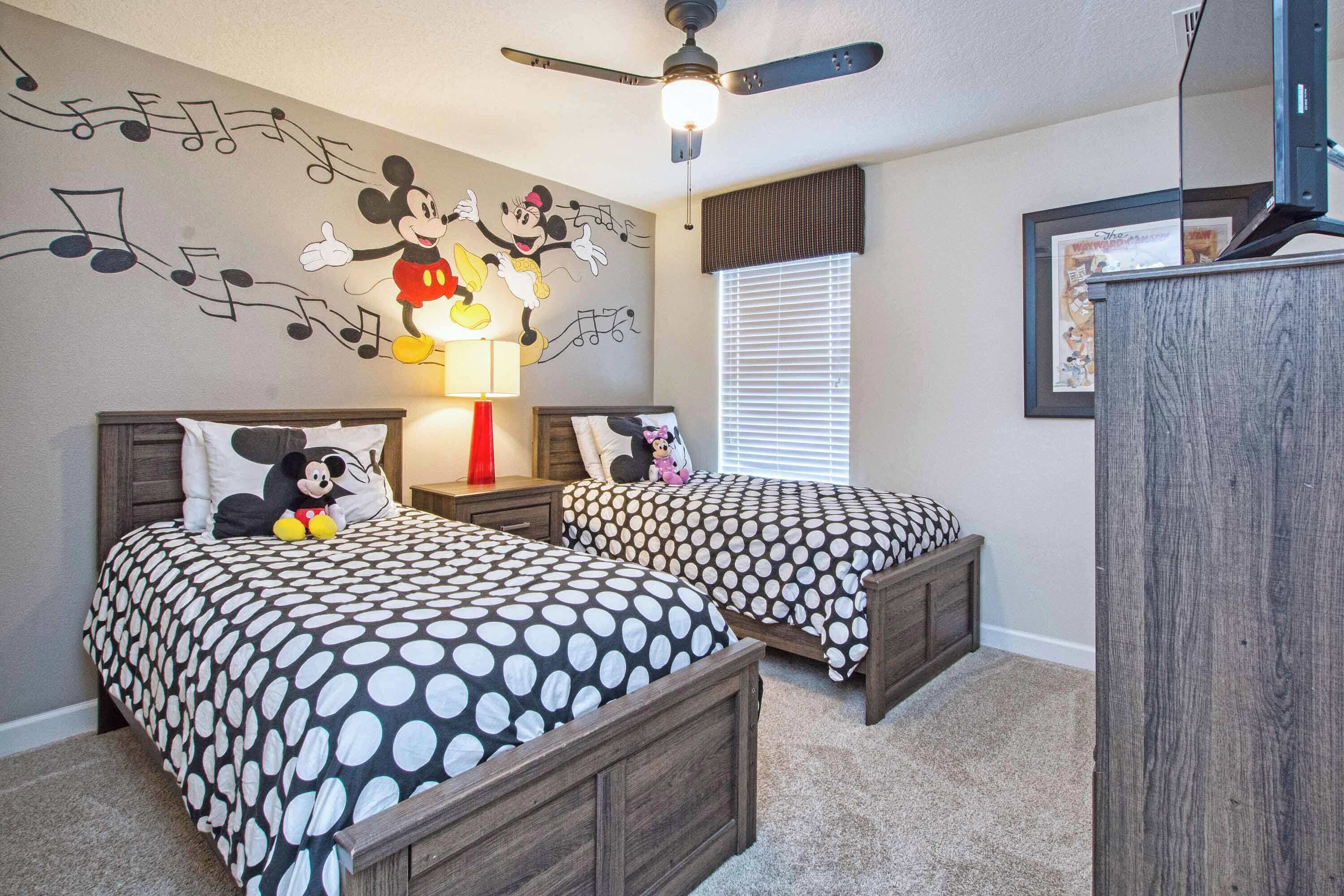 Une chambre avec deux lits, une fresque murale représentant Mickey Mouse, un ventilateur de plafond et une table de chevet avec une lampe.
