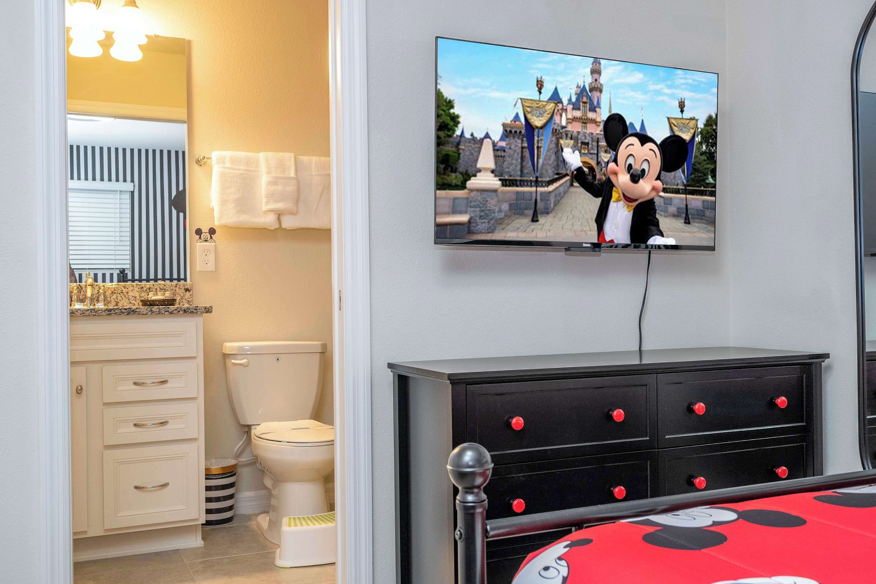 A bathroom with a toilet, a sink, a mirror, and a television displaying a cartoon character.
