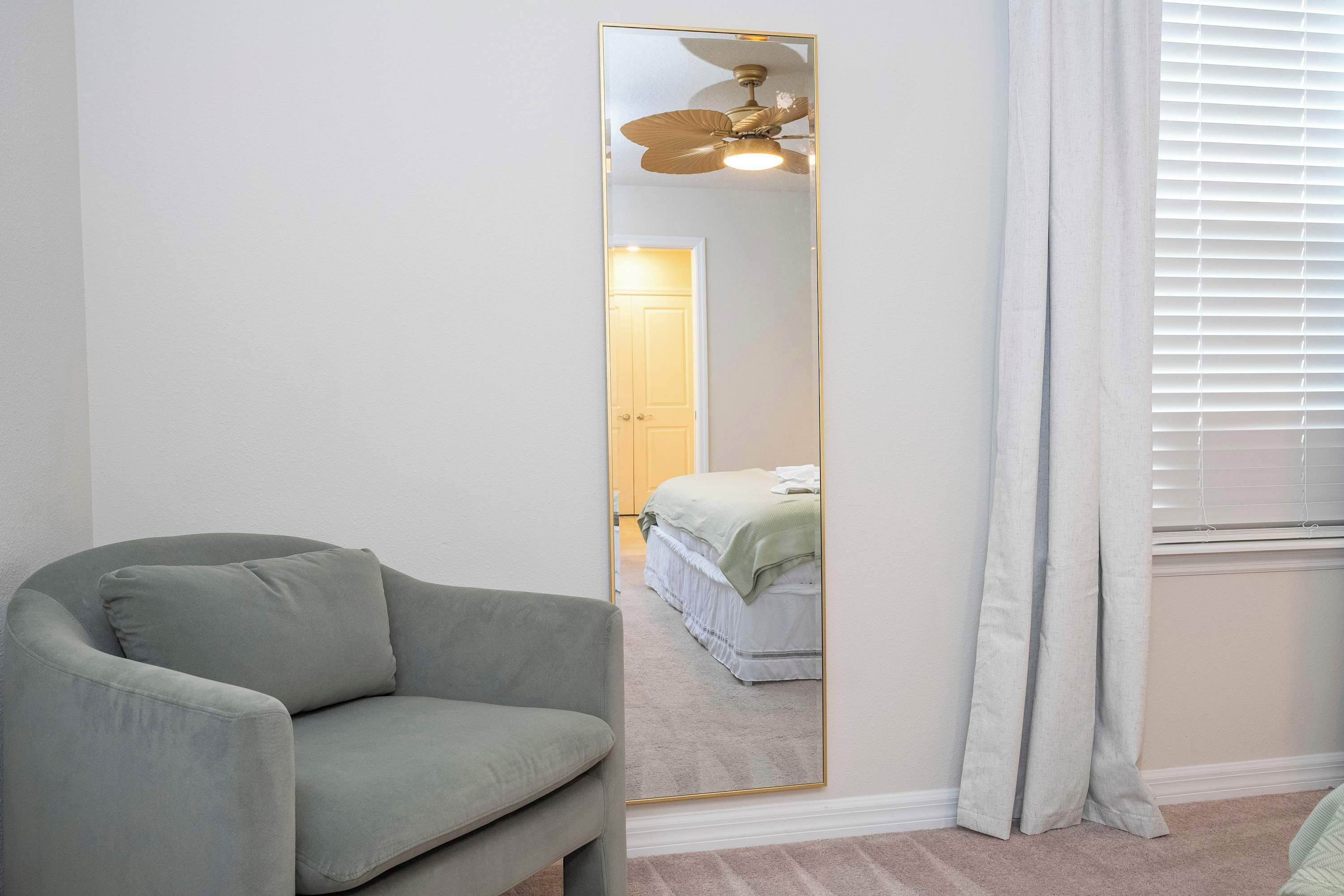 A room with a grey armchair, a full-length mirror, a ceiling fan, a bed with a green blanket, and a window with blinds.