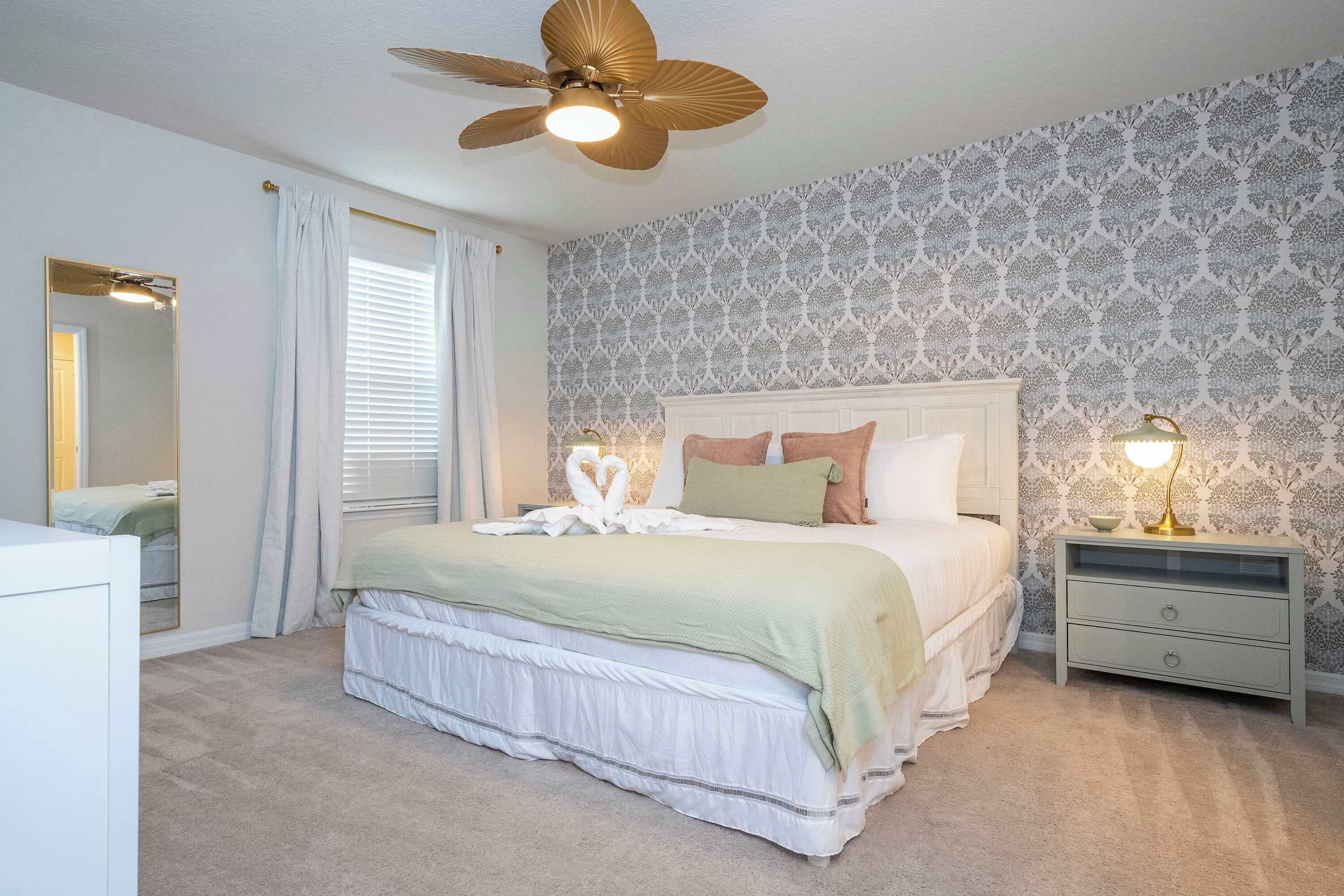 A bedroom with a bed, a nightstand, a ceiling fan, a mirror, and patterned wallpaper.