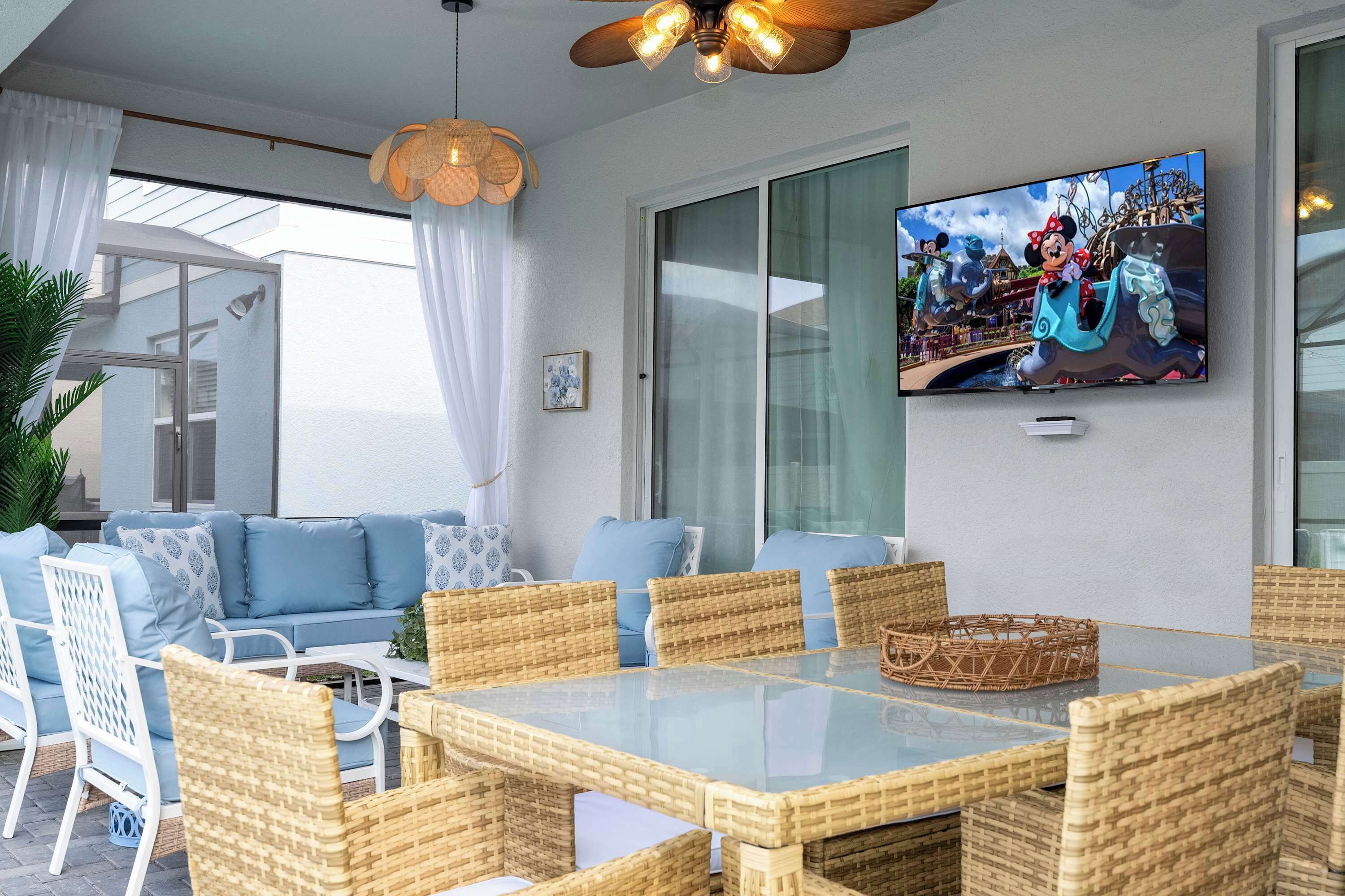 Un espace repas extérieur moderne, avec des meubles en osier, une table à plateau en verre et un téléviseur à écran plat fixé au mur.