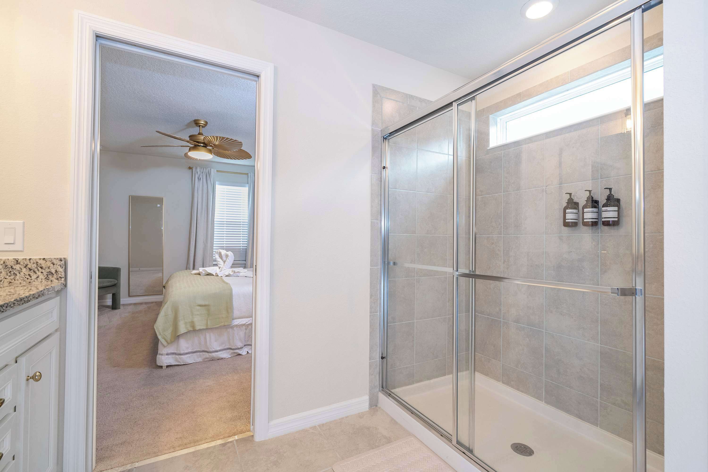 A bathroom with a glass shower enclosure, a ceiling fan, and a bed in the adjacent room.