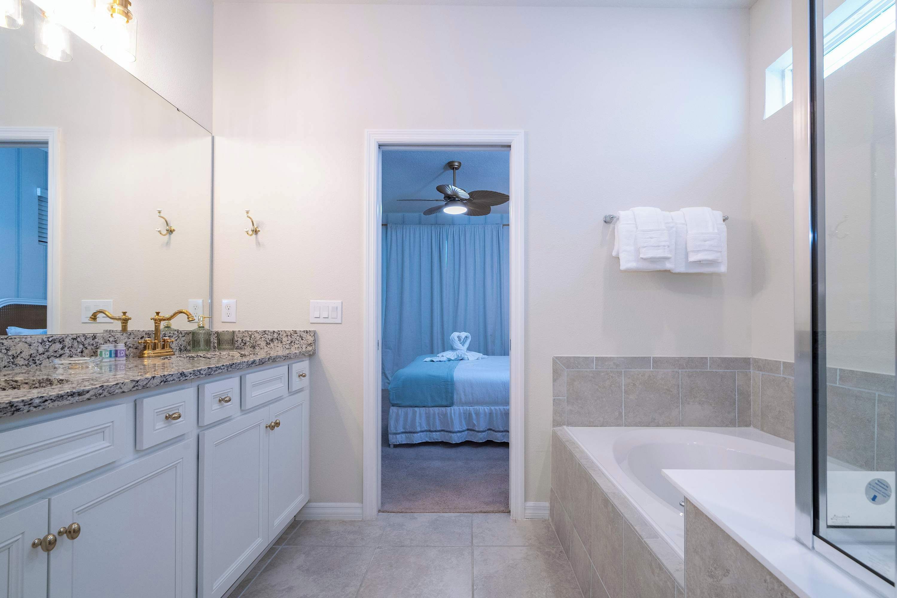 A bathroom with a bathtub, a shower, a vanity with a mirror, and a door leading to a bedroom with a bed and a ceiling fan.