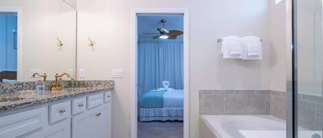 A bathroom with a bathtub, a shower, a vanity with a mirror, and a door leading to a bedroom with a bed and a ceiling fan.