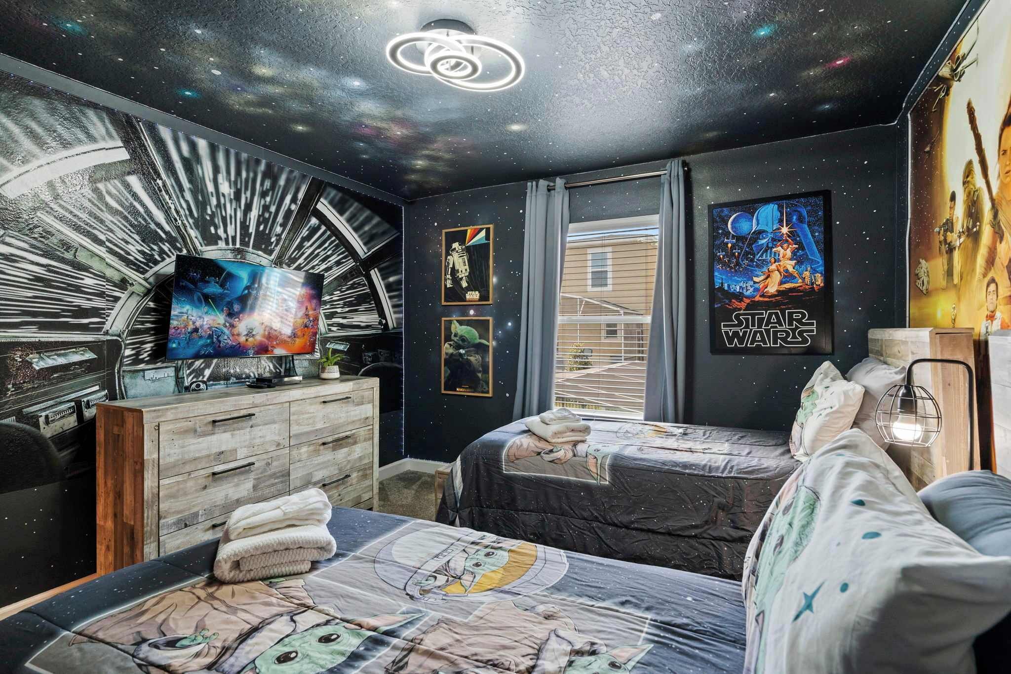 A room with a galaxy-themed wall, a Star Wars poster, a TV, and a bed with a space-themed bedspread.