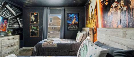 A bedroom with a starry ceiling, a bed with a galaxy-themed bedspread, a dresser, and a wall with Star Wars posters.