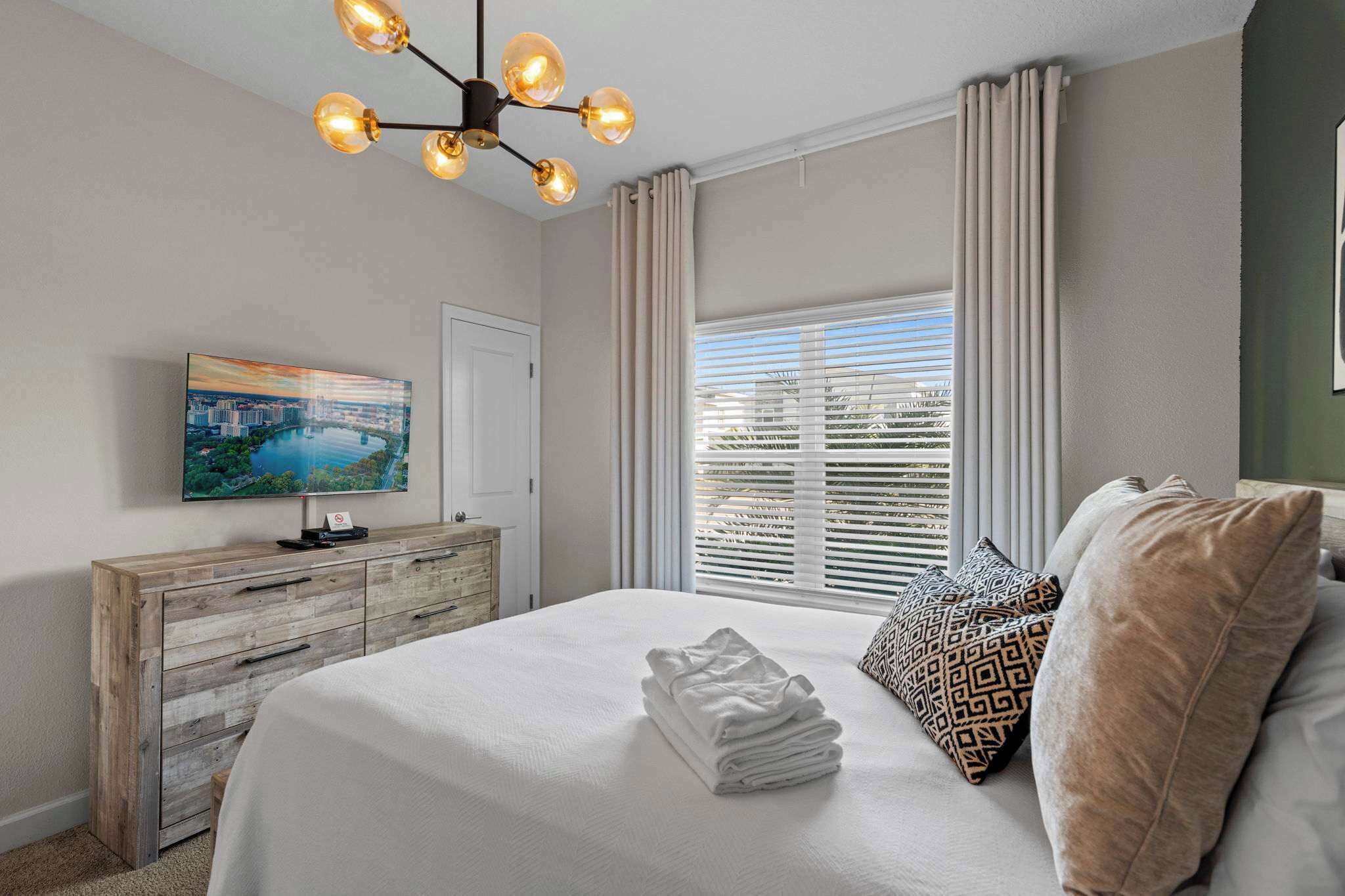 Une chambre d’hôtel comprenant un lit, une télévision, une commode en bois, une fenêtre avec des stores et un luminaire au plafond.