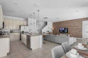 Una cocina moderna con armarios blancos, una isla central y un área de comedor con un sofá gris y un televisor montado en una pared de madera.