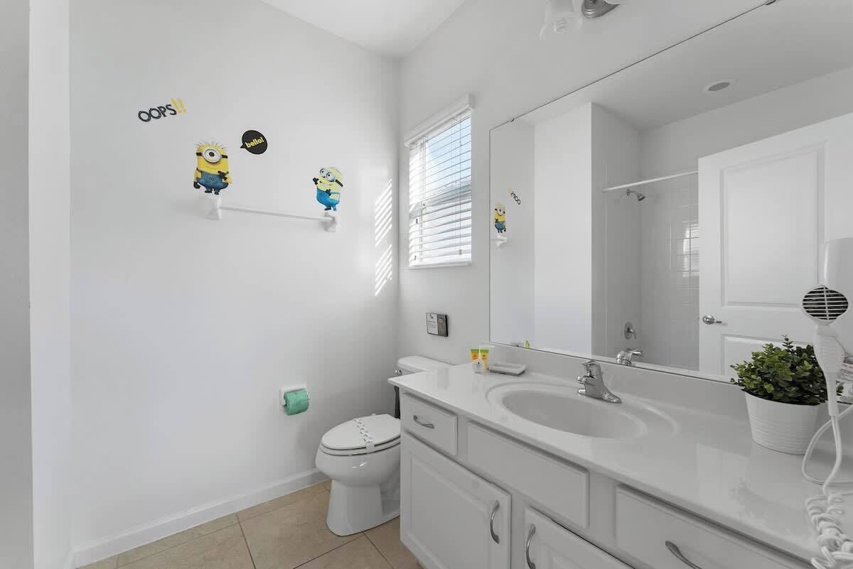 Un baño moderno con un lavabo, tocador y retrete blancos. Hay tres pegatinas de Minions en la pared, y un secador de pelo sobre el tocador.