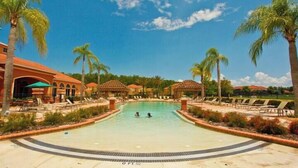 Piscina com duas pessoas na água, rodeada por palmeiras e cadeiras de descanso.