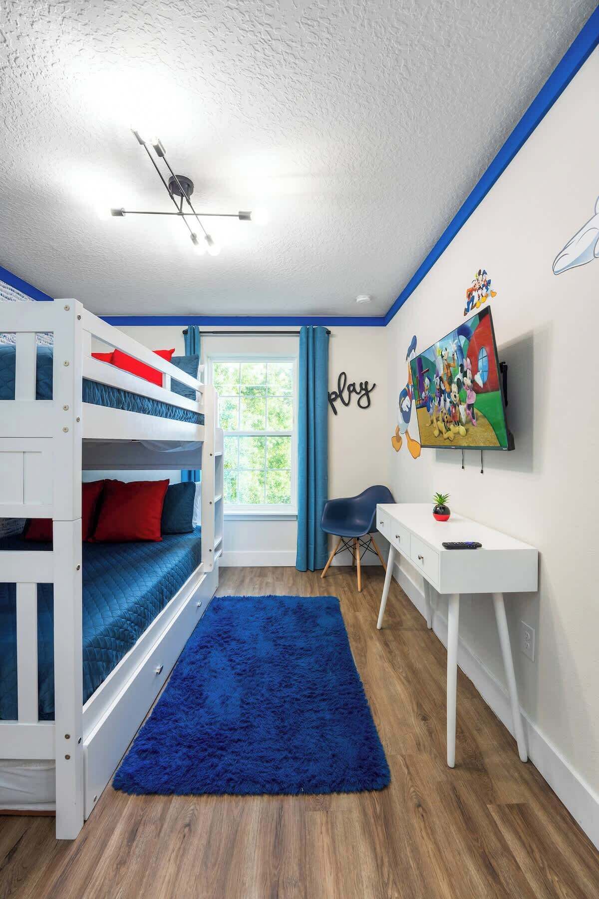 A child's bedroom with bunk beds, a desk, a TV, and a window with curtains.