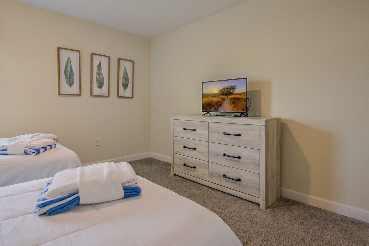 Ein Hotelzimmer mit zwei Betten, einem Schminktisch, einem Fernseher und gerahmten Kunstwerken an der Wand.
