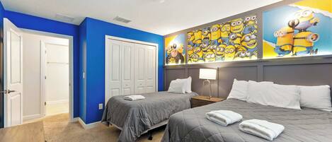 Habitación de hotel con dos camas, un escritorio, una lámpara y una pared decorada con un mural con tema de Minions.