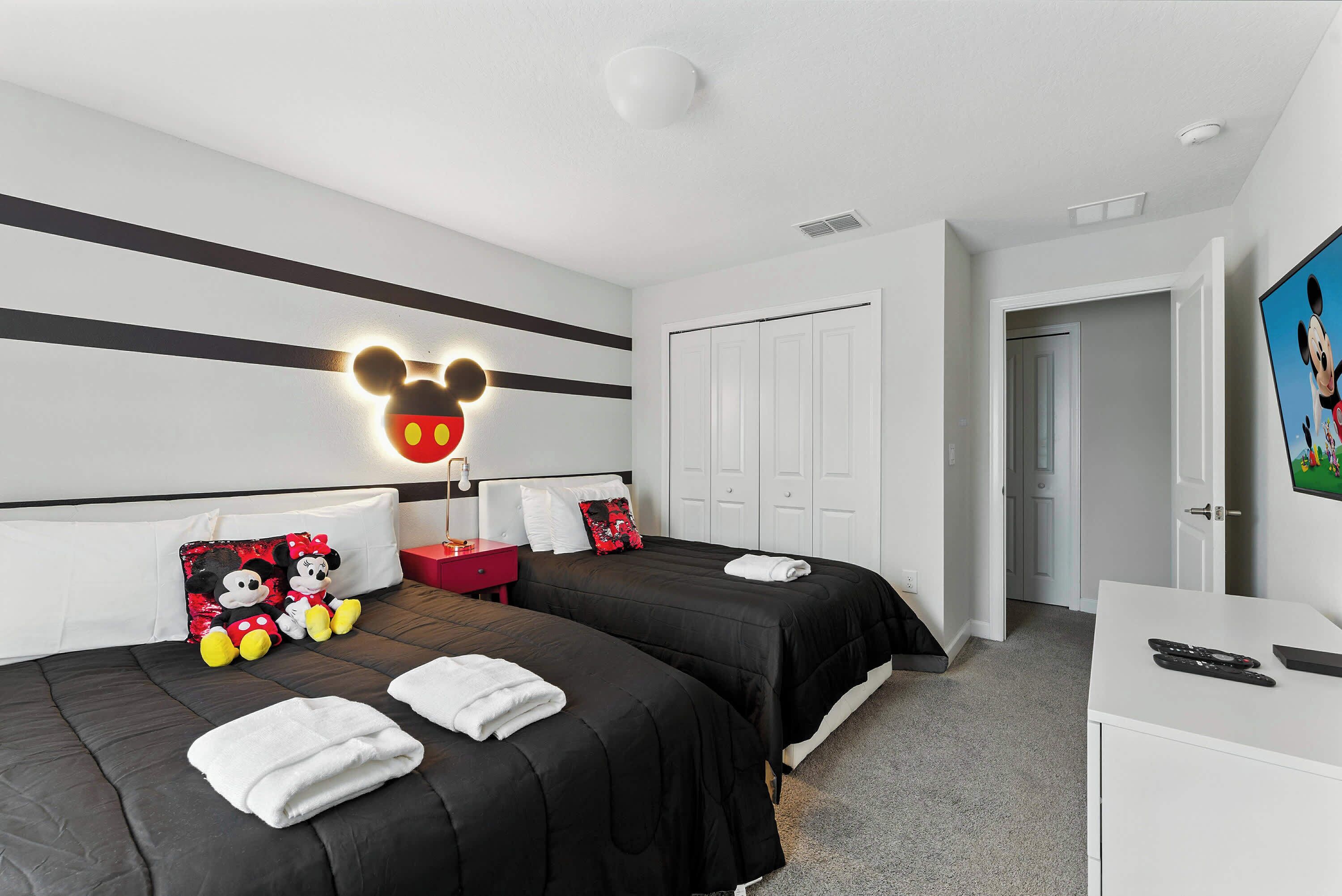 Ein Hotelzimmer mit zwei Betten, einer Mickey Mouse Wanddekoration, einem roten Nachttisch und einem weißen Schreibtisch mit Fernbedienung.
