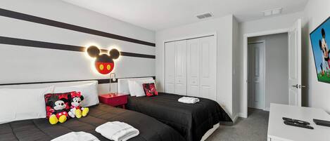 Ein Hotelzimmer mit zwei Betten, einer Mickey Mouse Wanddekoration, einem roten Nachttisch und einem weißen Schreibtisch mit Fernbedienung.