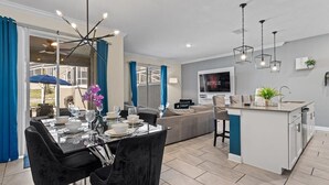 A modern living room with a dining area, a kitchen island, and a TV mounted on the wall. - Champions Gate Oasis, 4BR Vacation Haven (Davenport)