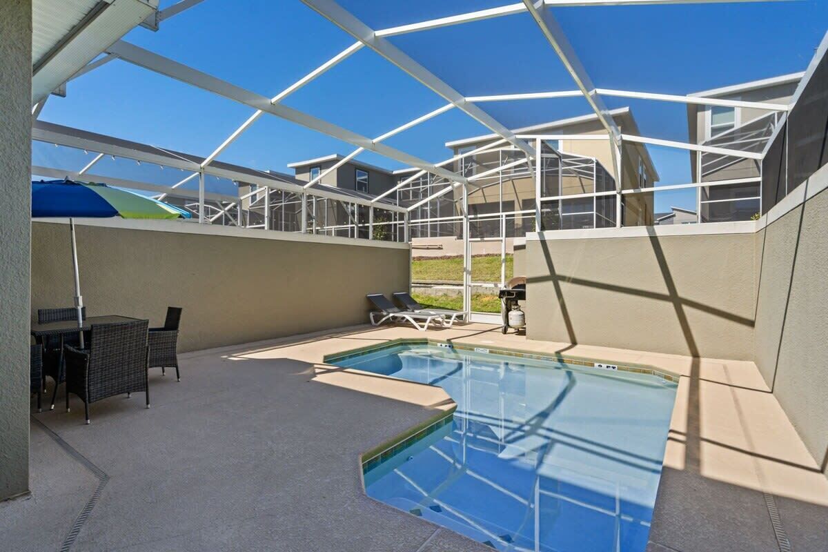 Una piscina con cerramiento de vidrio, sillas de descanso y una mesa con sillas.