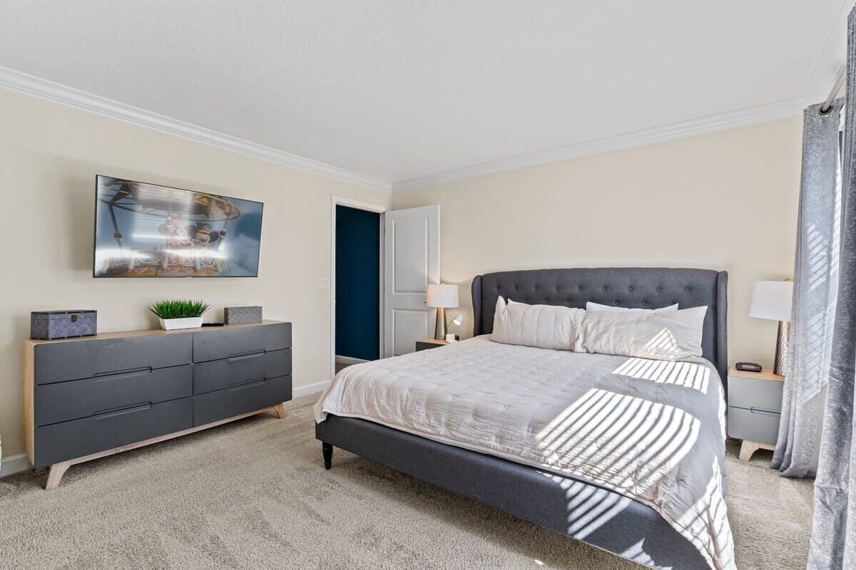 Une chambre à coucher comprenant un lit, une commode, une table de chevet, une lampe, un tableau et une porte.