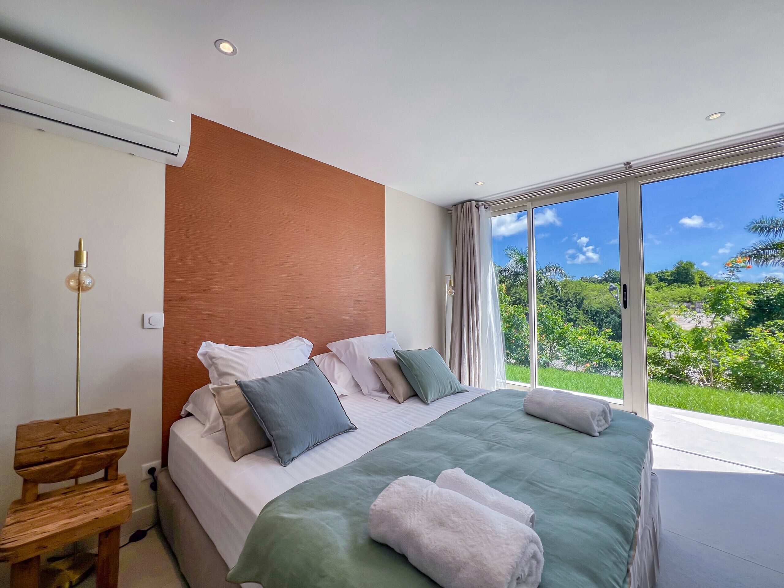 A modern bedroom with a large bed, a wooden bench, and a view of greenery through large windows.