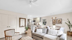 A modern living room with a sofa, ottoman, and a wooden chair. There is a ceiling fan, a kitchen area in the background, and a dining table with chairs. - Beachwood Villas 1G - The Nest at Seagrove (Santa Rosa Beach)
