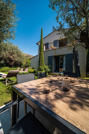 Uma mesa de madeira com cadeiras e uma garrafa de água em um ambiente externo ensolarado, com uma casa ao fundo.