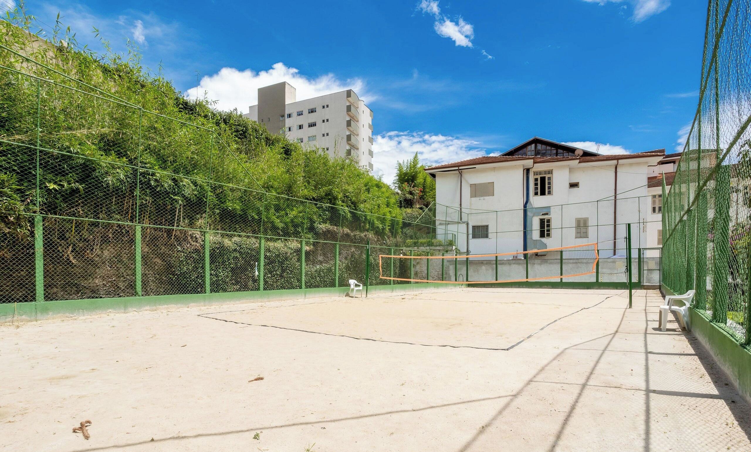 An outdoor tennis court with a net, surrounded by a green fence, and adjacent to a white building with a red roof.