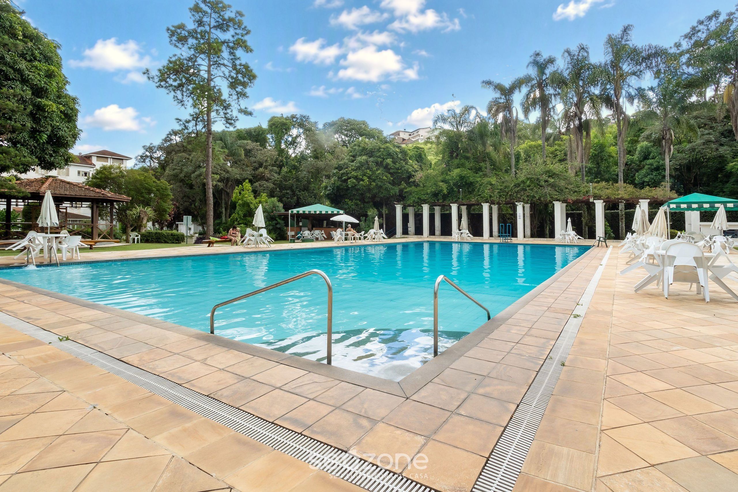 A swimming pool with a tiled deck, surrounded by trees and shaded areas with umbrellas.