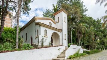 Um edifício branco com um campanário, rodeado por vegetação e um caminho de pedras.