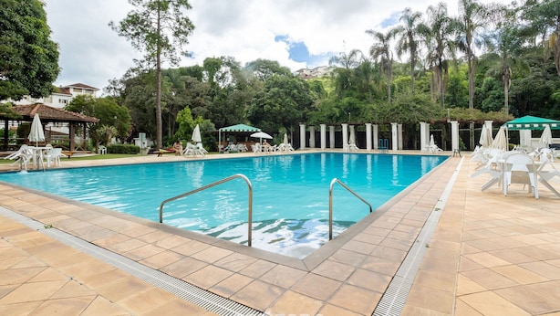 Piscina com bordas azulejadas, rodeada por cadeiras de descanso e guarda-sóis, em área ajardinada com árvores e um gazebo.