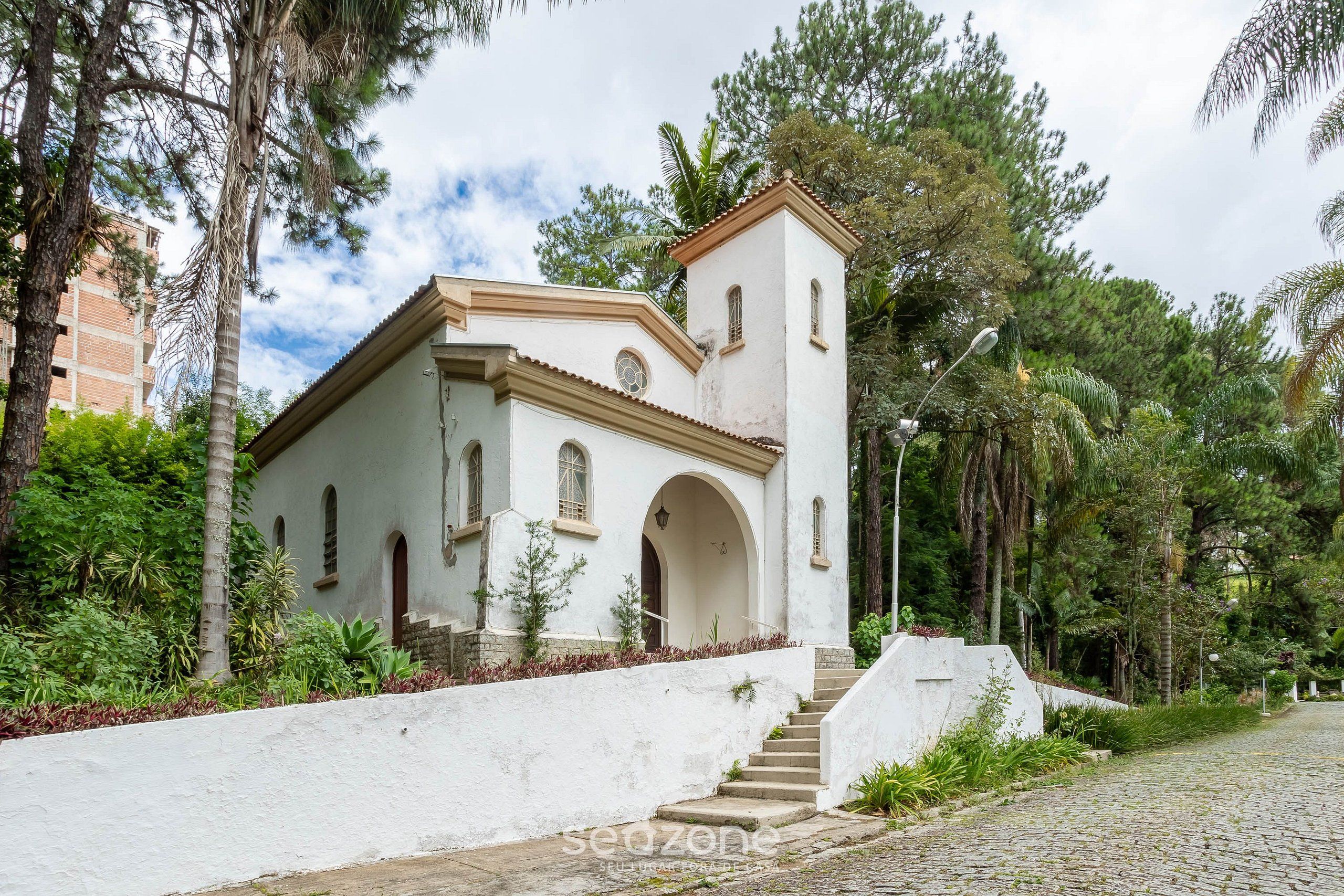 Um edifício branco com um campanário, rodeado por vegetação e um caminho de pedras.