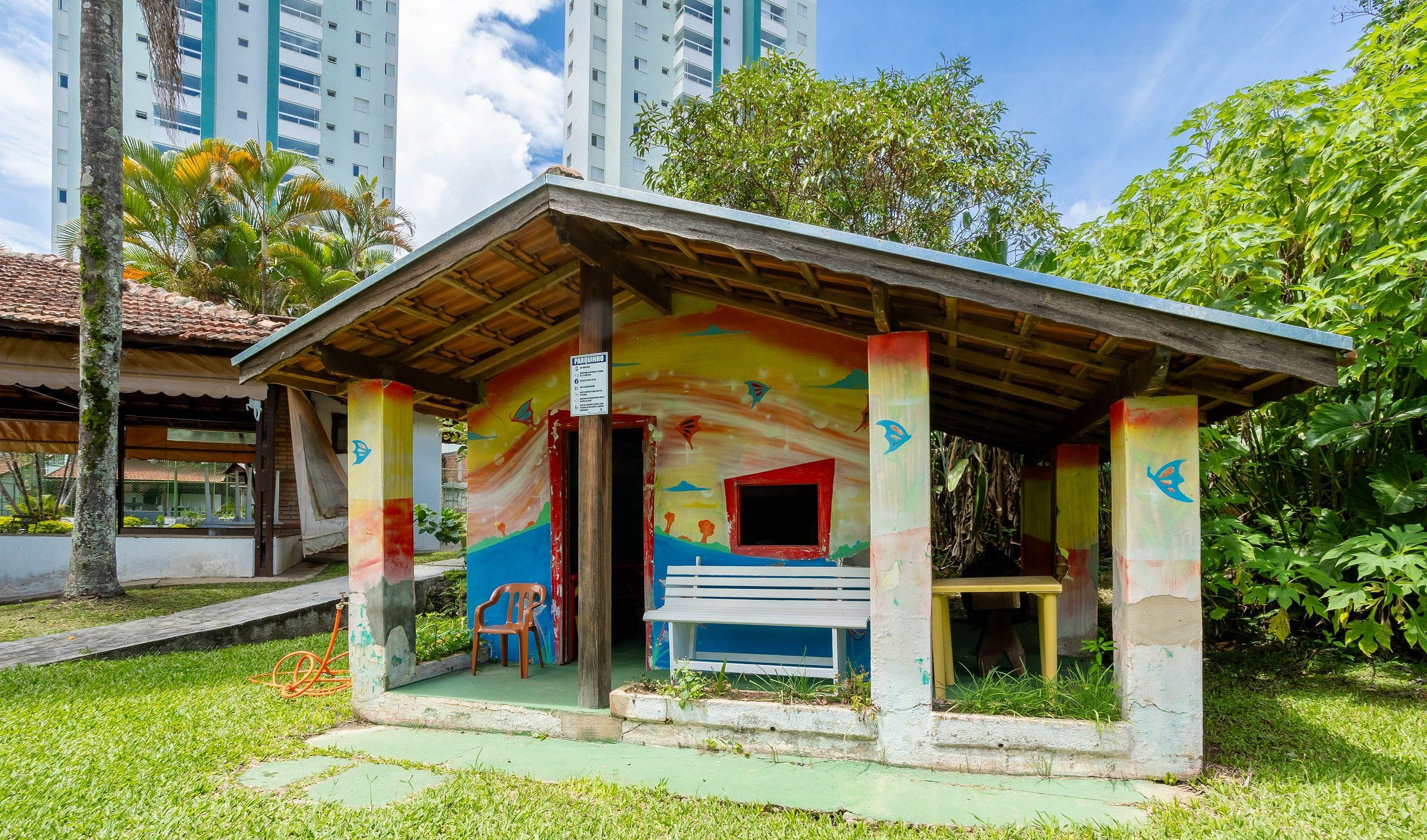 Un pequeño pabellón colorido con techo inclinado, rodeado de abundante vegetación y altos edificios al fondo.