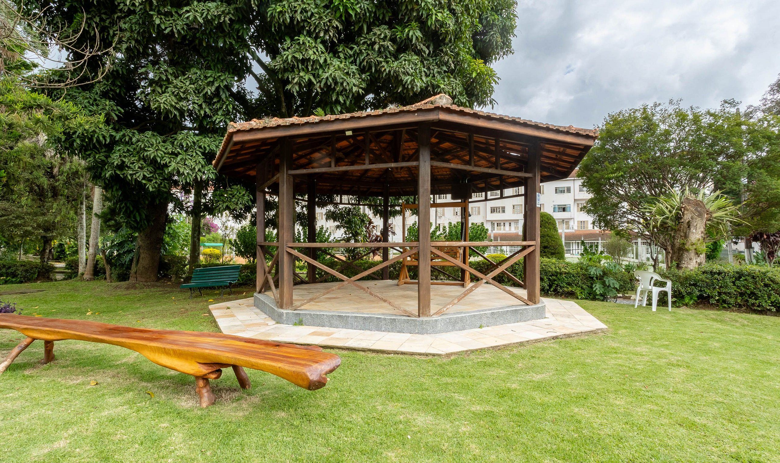 A wooden gazebo with a tiled floor, surrounded by grass and trees, with a bench and a playground slide nearby.