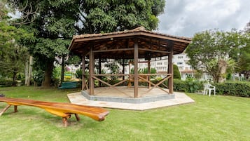Un pabellón de madera con suelo de baldosas, rodeado de césped y árboles, con un banco y un tobogán cerca.