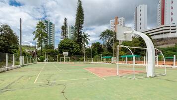 Una cancha de baloncesto al aire libre con un aro, rodeada por una valla, árboles y edificios altos al fondo.