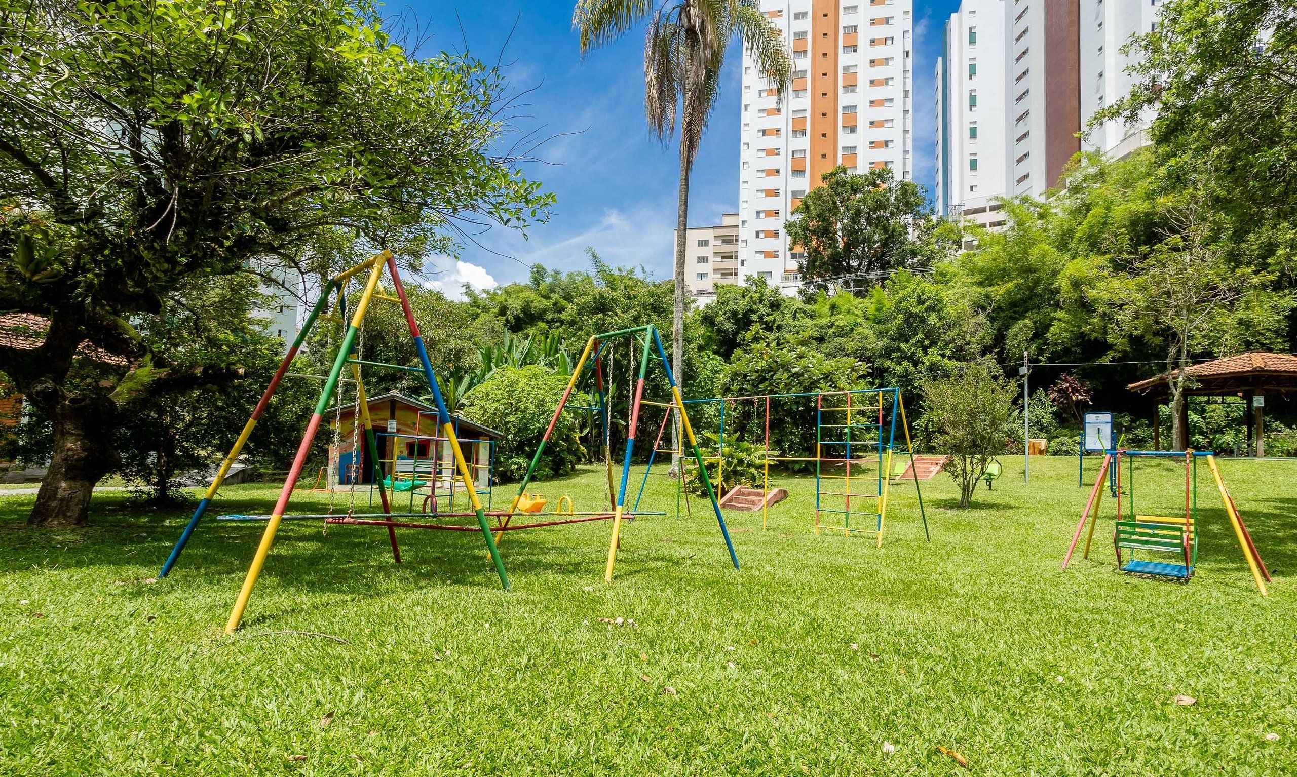 Um parquinho com balanços e estruturas de escalada, rodeado por grama e árvores, com edifícios altos ao fundo.