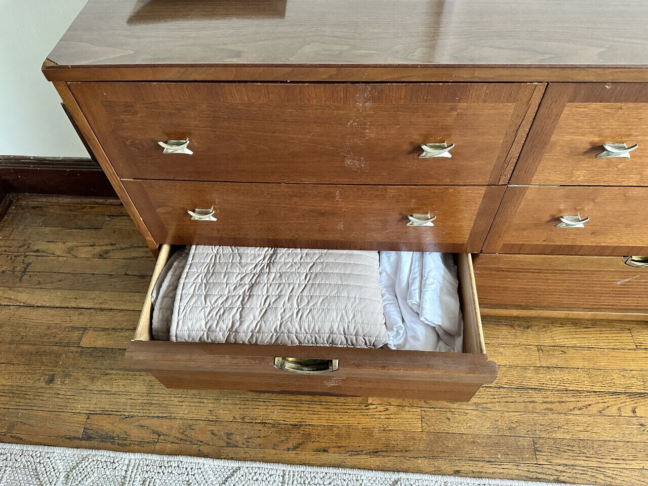 Une commode en bois avec des tiroirs, dont un est ouvert et laisse apparaître un lit recouvert d’une couverture matelassée et de draps blancs.