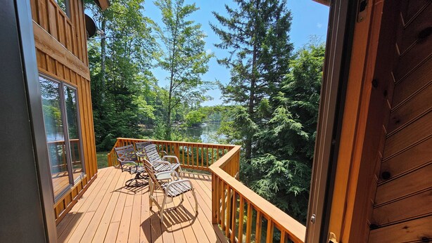 Terraza de madera con dos sillas y una mesa, con vistas a un bosque y un lago.