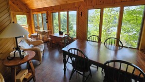 Interior de una cabaña de madera con una mesa de comedor, sillas, una lámpara y vista a un lago.