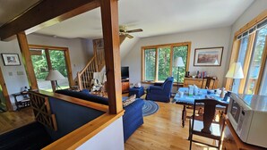 Amplia sala de estar con escalera de madera, comedor con mesa y sillas, y ventanas grandes que ofrecen vistas al exterior.