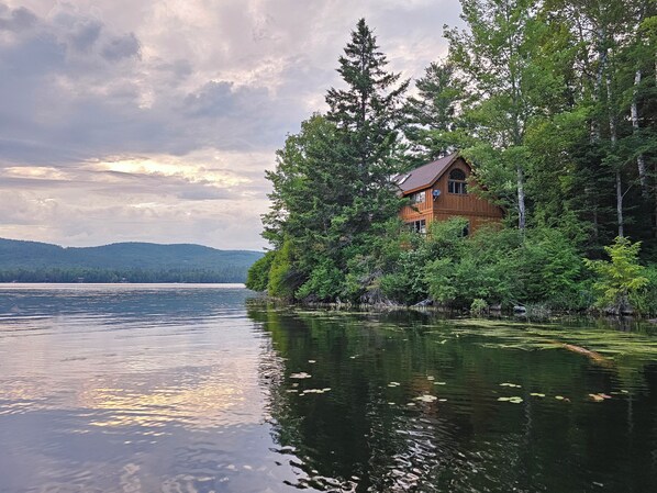 Una cabaña rodeada de un bosque frondoso, un lago y una cadena montañosa distante.