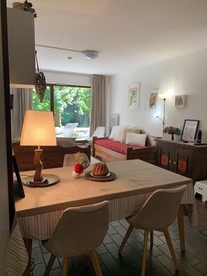 A room with a dining table, two chairs, a bed with a red blanket, a bedside table with a lamp, and a view of greenery outside.