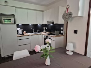 A modern kitchen with white cabinets, a black backsplash, a microwave, a sink, and a dining area with a vase of flowers.