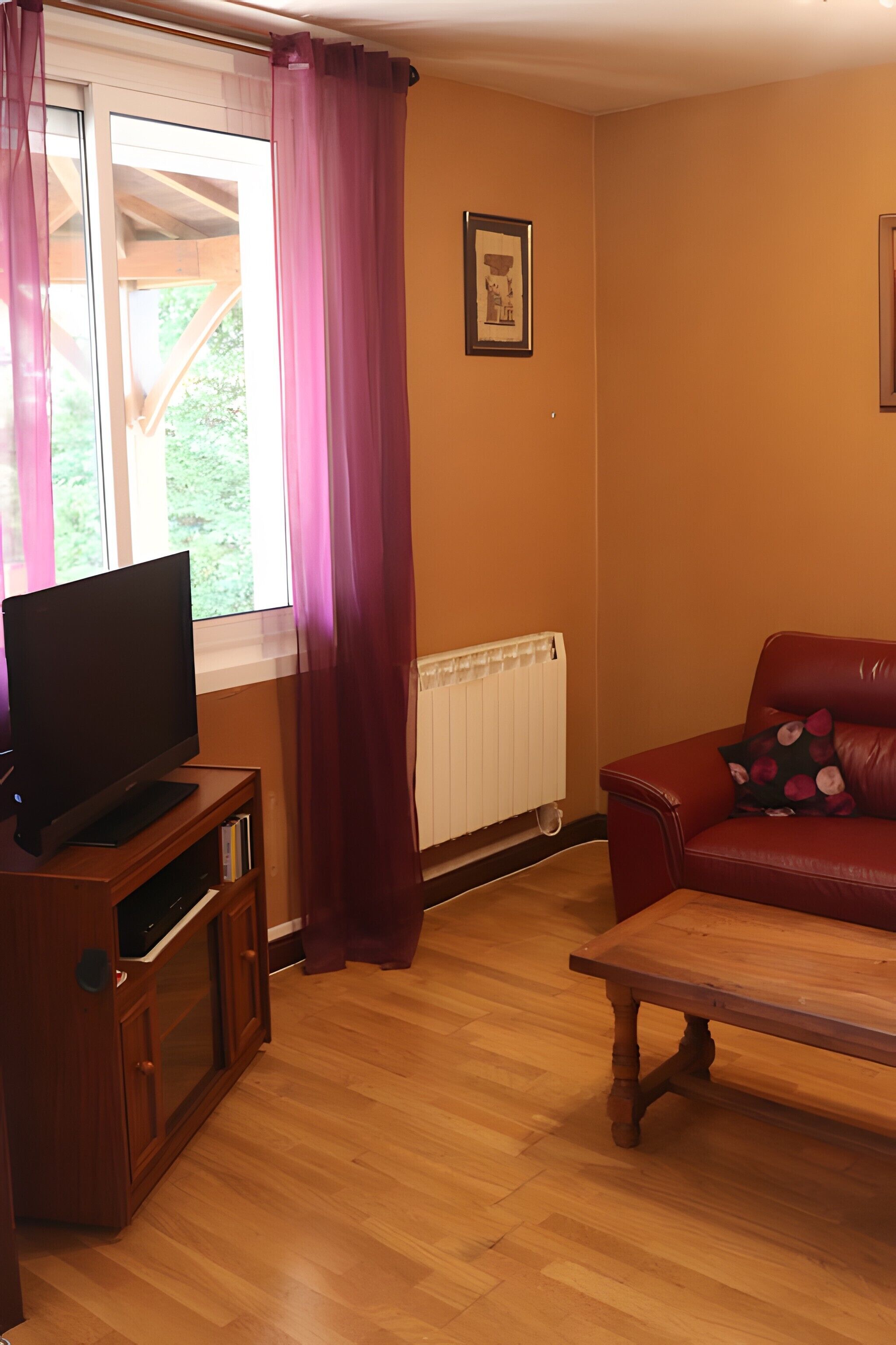 A living room with a television on a wooden stand, a red leather sofa, a wooden coffee table, and a window with curtains.