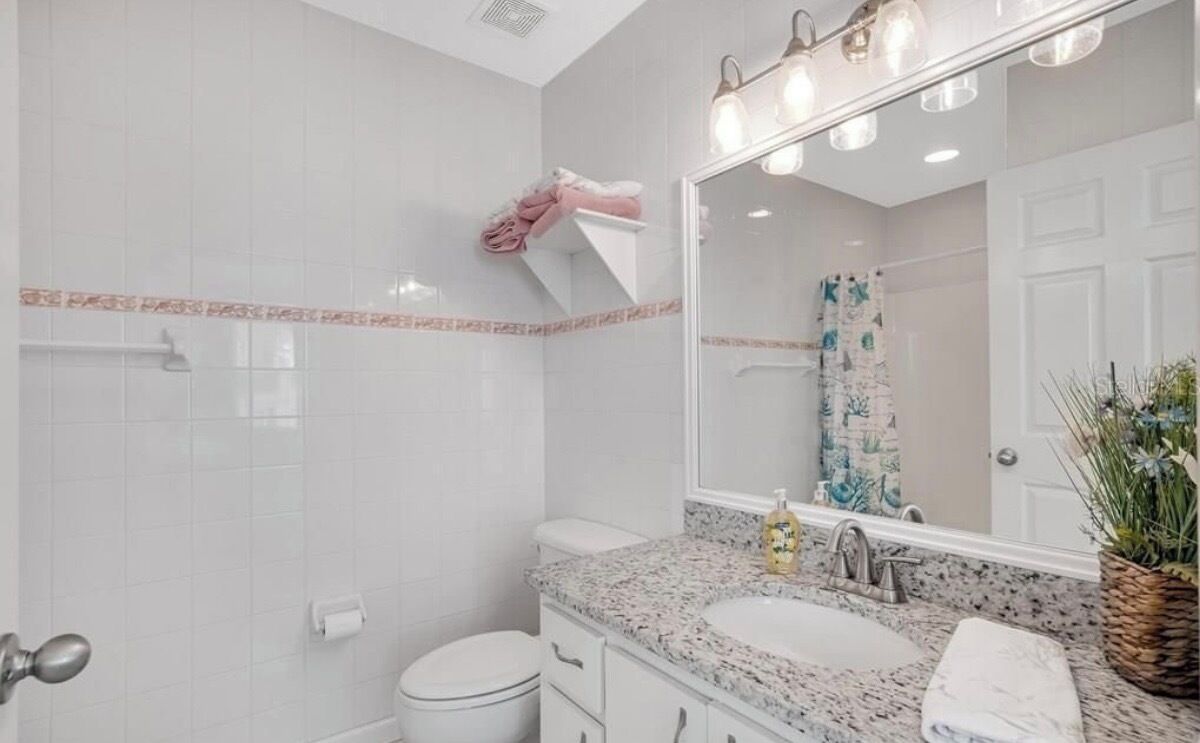 Un baño con azulejos blancos, un lavabo con encimera de mármol, un espejo con iluminación, un perchero para toallas y una planta.