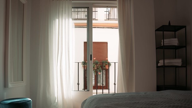 Una camera da letto con un letto, una finestra con tende, un balcone con piante e uno scaffale con asciugamani.