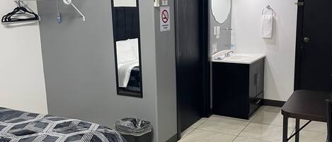 A hotel room with a bed, a bathroom with a sink and mirror, a towel rack, and a trash bin.