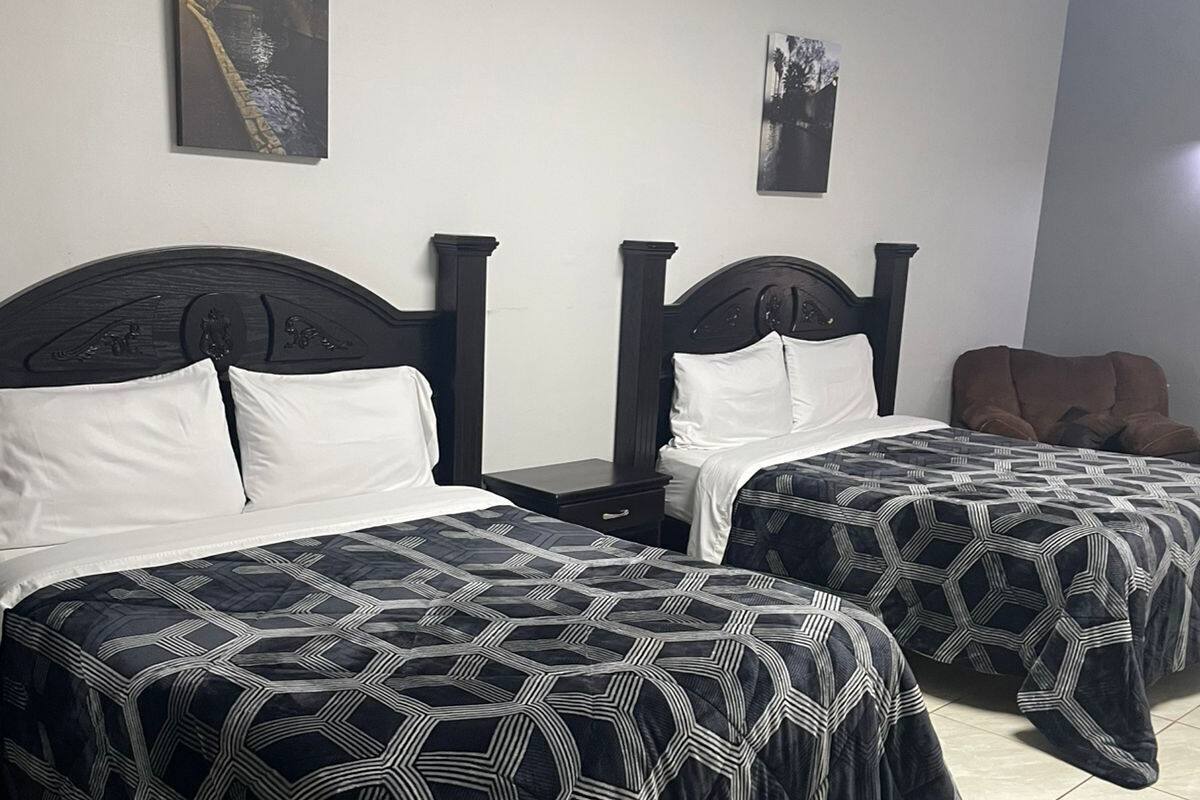 Habitación de hotel con dos camas, cada una con cabecero oscuro y ropa de cama estampada. Hay dos mesitas de noche, una silla marrón y obras de arte en las paredes.