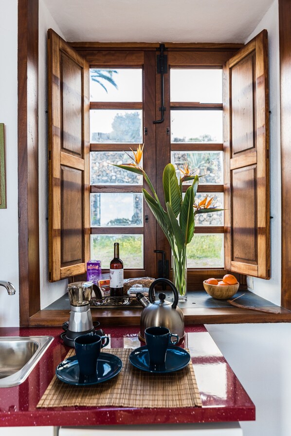 A kitchen with wooden cabinets, a window with open shutters, a coffee maker, a kettle, and a bowl of fruit on the windowsill.