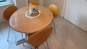 Spiseområde med et rundt bord, fire stole, en hylde med køkkenudstyr, et vindue og en dør.